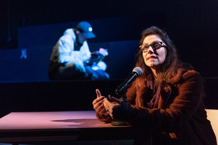 A woman wearing and winter coat and glasses is sitting at a table. She is speaking into a microphone.