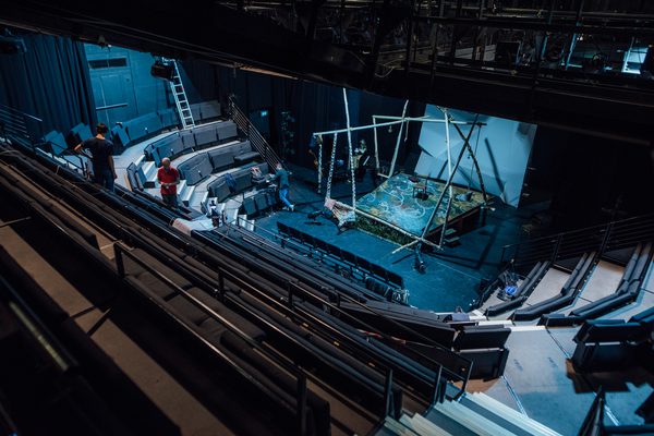 Image of Traverse 1 from top right-hand side steps. Two people stand on the left-side steps. a third person is on stage level leaning against a metal bar above audience seating in the front row, left side. The stage set is made of several wooden polls connecting on angles, with a white floral painted raised floor, a wooden chair centre stage, a bucket hanging from a rope on the right, a cushion and blanket on the front left corner of the set, and grass along the front of the raised floor.