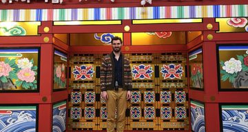 Andrew Thompson, a tall, dark haired and bearded man, stands in front of an elaborately painted doorway. The door are painted with vivid tiles with images of flowers.