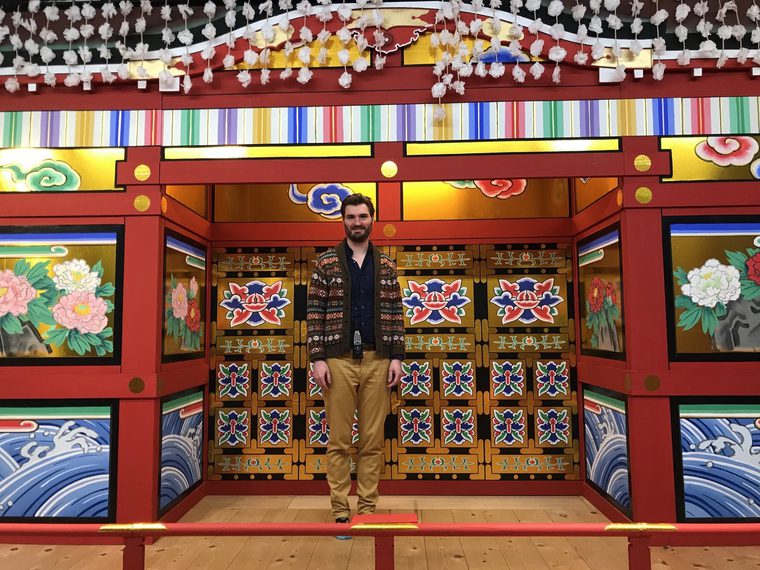 Andrew Thompson, a tall, dark haired and bearded man, stands in front of an elaborately painted doorway. The door are painted with vivid tiles with images of flowers.