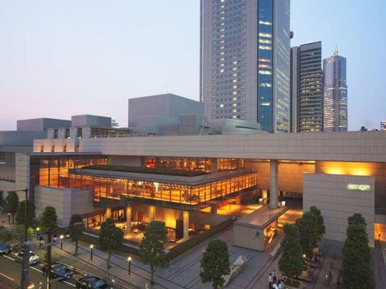 The National Theatre of Japan: a large, rectangular, modern looking building made of white stone with four stories. It is fronted by an avenue of tall trees and lit by internal orange lights.