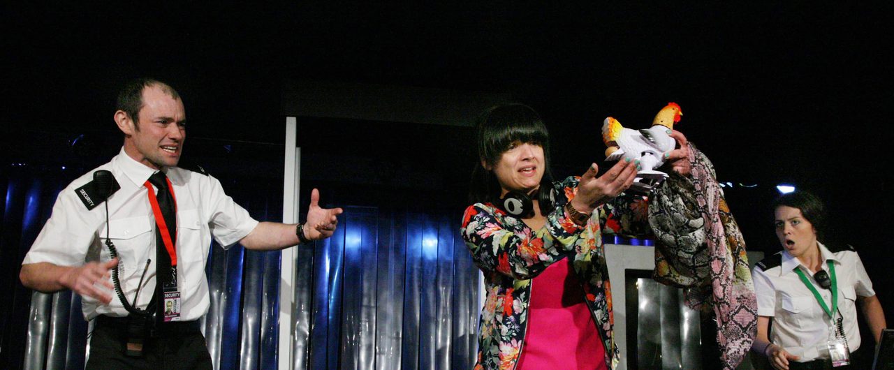 A woman holds a toy plastic chicken. Two airport security guards behind her look angry.