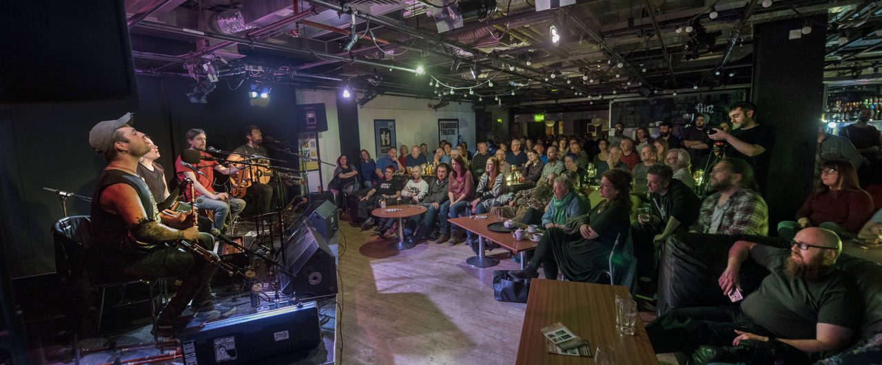 Gig performance in the Traverse bar. Four musicians- a guitarist, fiddler, concerntinist, and bagpiper sit on the corner stage in front of microphones singing. To the right a full audience sits and stands listening to the music.