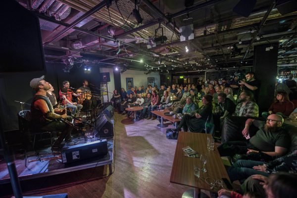 Gig performance in the Traverse bar. Four musicians- a guitarist, fiddler, concerntinist, and bagpiper sit on the corner stage in front of microphones singing. To the right a full audience sits and stands listening to the music.